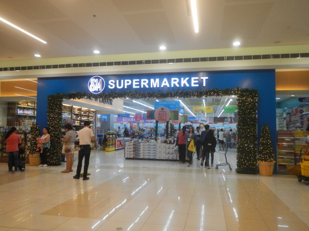 Eine Gruppe von Menschen, die vor einem Supermarkt stehen, einige halten Einkaufstaschen, mit einem Einkaufswagen, einem Topf mit einer Pflanze, Regalen mit Gegenständen, Schildern und einer Decke mit Leuchten.