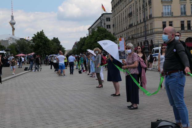 Eine Gruppe von Menschen marschiert auf einer Straße in Berlin und hält Schilder und Banner hoch, einige tragen Masken und Taschen oder Schirme, während im Hintergrund Bäume, Gebäude, geparkte Fahrzeuge und ein turmförmiger Fahnenmast zu sehen sind.