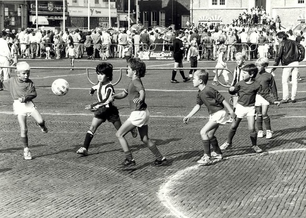 Schwarzes Bild von jungen Jungs, die auf der Straße Fussball spielen, umgeben von einer Zuschauermenge hinter einem Geländer, Gebäuden und Polen im Hintergrund.