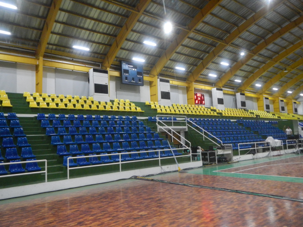 Eine geräumige Indoor-Sporthalle mit einem Holzboden, blauen und gelben Stühlen, Geländern, Treppen zum Sitzbereich, wandmontierten Bildschirmen, Deckenleuchten und einigen anwesenden Personen.