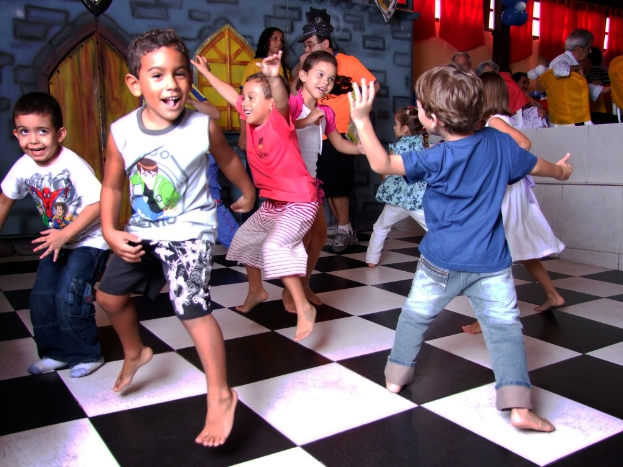 Eine Gruppe von Kindern, die auf einem Boden tanzen, mit Menschen, Stühlen, Wänden, Kleidung und verschiedenen Gegenständen im Hintergrund.