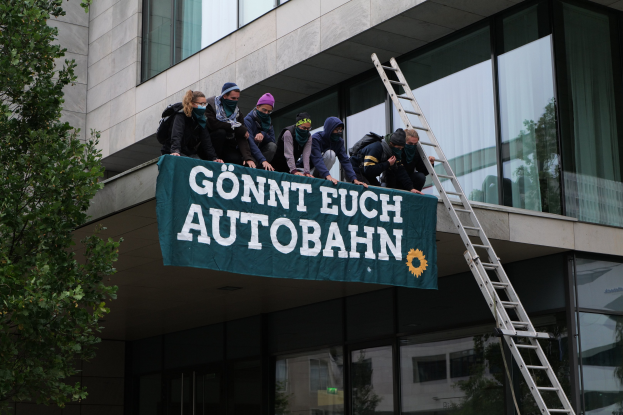 Eine Gruppe von Menschen auf einem Gebäude mit einer Fahne, mit einem Baum links und Glasfenstern im Hintergrund, wahrscheinlich protestierend für die deutsche Regierung.
