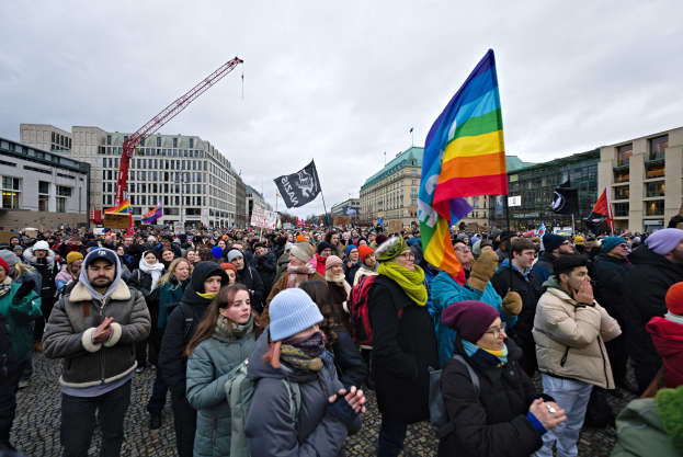 Eine große Gruppe von Menschen steht vor einem Gebäude und hält Fahnen und Schilder mit der Aufschrift "Lgbtq+ rights march in Berlin", einige tragen Mützen und Taschen, im Hintergrund sind Gebäude, ein Kran und ein bewölkter Himmel zu sehen.