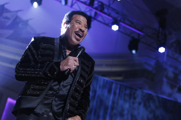Lionel Richie performs bei den Grammys in einem schwarzen Kleid und hält ein Mikrofon auf einer hell erleuchteten Bühne.