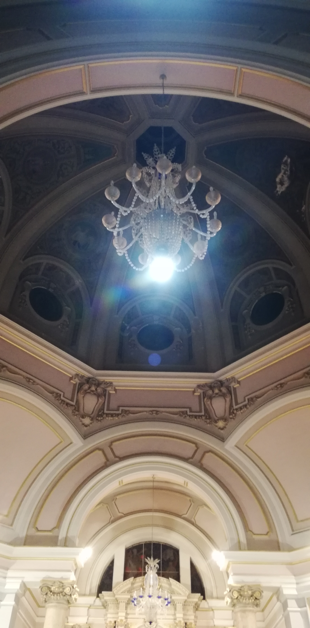 Ein großer Kronleuchter hängt von der Decke und erhellt einen Raum mit detaillierten Designs und Lichtern, während Skulpturen die Wände schmücken.