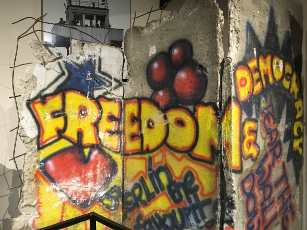 Mauer mit Graffiti, das "Freiheit und Demokratie" in fetten schwarzen Buchstaben liest, einem Geländer am Boden und einem Gebäude mit Fenstern im Hintergrund.