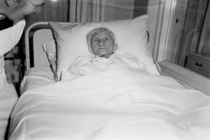 Eine ältere Frau liegt in einem Krankenhausbett mit einem weißen Laken und Kissen, während eine Krankenschwester neben ihr steht, mit einem Fenster und Vorhängen im Hintergrund.