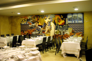 Ein Restaurant-Innenraum mit für ein Essen gedeckten Tischen, mit Tellern, Gläsern, Besteck und Servietten, einem Bild an der Wand und Deckenlampen, die eine gemütliche Atmosphäre schaffen.