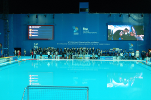 Gruppe von Menschen, die um ein Hallenbad mit Netz am Boden stehen, mit einer Wand im Hintergrund, die den Text und Logos "FINA World Championships 2015 Kazan" anzeigt, sowie Gerüste und Lampen.