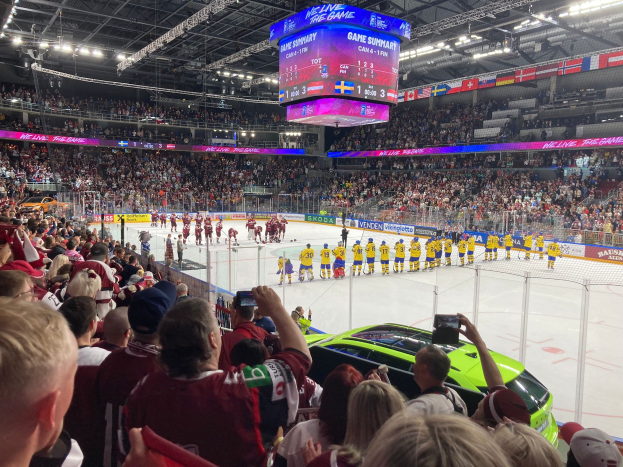 Ein Eishockeyspiel in einer großen Arena mit Spielern auf dem Eis, Zuschauern auf Stühlen, Fotografen mit Kameras, Arenabeleuchtung und einem Bildschirm mit Text über dem Eis.