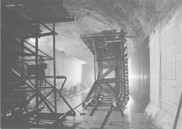 Schwarzes und weißes Foto eines Tunnels mit einer Metallstruktur in der Mitte, Metallstangen auf der linken Seite, einer Wand auf der rechten Seite und Text unten.