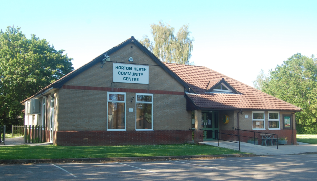 Ein Haus mit Dach, Fenstern, Türen, einem Schild mit der Aufschrift "Horton Heath Community Centre", einem Metallzaun, einer Bank, Topfpflanzen, Gras, einem Weg, einer Baumgruppe und einem bewölkten Himmel.
