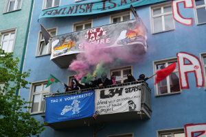 Eine Gruppe von Menschen auf einem Balkon mit Fahnen und Transparenten, mit einem Baum links und Rauch in der Luft, während einer Demonstration.