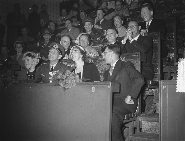 Schwarzes und weißes Foto einer Menschenmenge in einem Stadion mit einem Podium und einem Blumenstrauß im Vordergrund, Stufen auf der rechten Seite und einer Wand im Hintergrund.