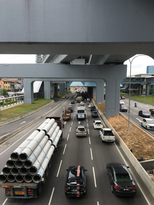 Eine vielbefahrene Autobahn mit mehreren Fahrzeugen, umgeben von einer städtischen Landschaft mit einer Brücke, Straßenlaternen, Gras, Gebäuden, Bäumen und dem Himmel.