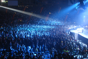 Eine große Menschenmenge in einem hell erleuchteten Konzertsaal schaut Musikern auf der Bühne zu, die hinter Gittern spielen.