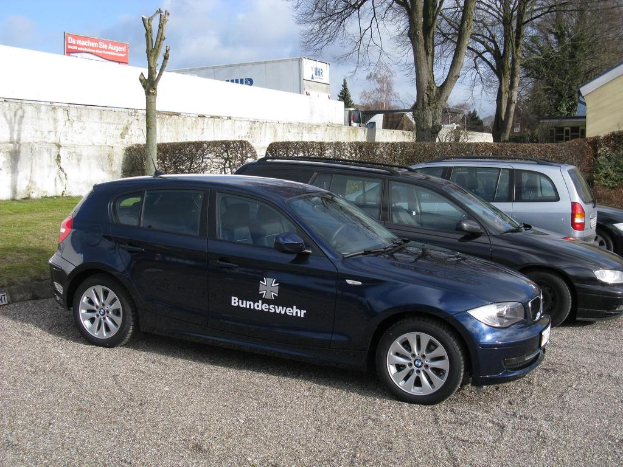 Mehrere Autos stehen auf einem von Gras, Büschen und Bäumen umgebenen Weg. Im Hintergrund sind Gebäude, ein Schild mit Text und ein Logo zu sehen.