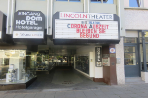Das Lincoln Theater in Berlin, Deutschland, ein Gebäude mit Glasfenstern und -türen und einer Tafel mit Text darauf, das verschiedene Objekte zeigt und den Eindruck einer pulsierenden Stadtlandschaft vermittelt.