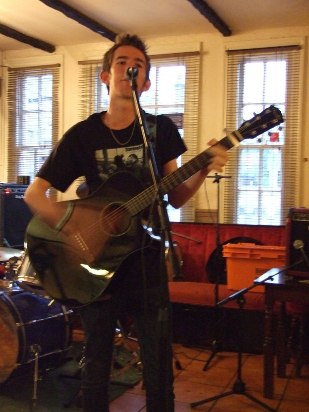 Ein Mann in einem schwarzen T-Shirt und Hose steht mit einer Gitarre und einem Mikrofon auf einem Ständer, mit einer blauen Trommel in der linken Ecke, einer Couch mit Gegenständen darauf und durchsichtigen Vorhängen im Hintergrund.