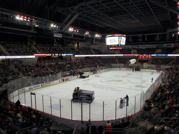 Ein Eishockeyspiel in einer großen Arena mit Fahrzeugen auf dem Eis, Zuschauern um die Bahn und Menschen auf den Tribünen, sowie Brettern, Lampen und einem Bildschirm im Hintergrund.