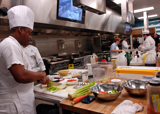 Eine Gruppe von Köchen in einer Küche beim Zubereiten von Speisen auf einem Schneidbrett, mit einem Koch im Vordergrund in einer weißen Schürze und Mütze, der ein Messer hält und von Utensilien, Schüsseln, Gemüse, einem Mobiltelefon, Flaschen und anderen Gegenständen umgeben ist, mit einem Fernseher und Deckenlampen im Hintergrund.