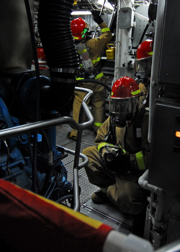Feuerwehrleute in Helmen und Schutzausrüstung arbeiten an einem Feuerwehrauto mit Rohren und anderen Gegenständen im Hintergrund, unter einem Licht an der Spitze.