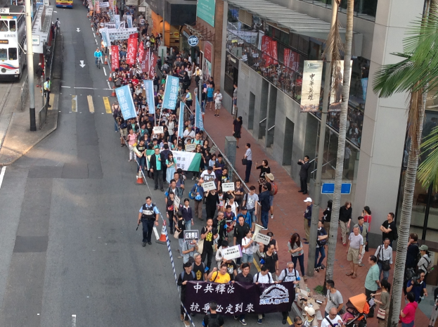 Eine große Gruppe von Menschen marschiert auf einer Stadtstraße mit Schildern und Transparenten, mit Fahrzeugen, Bäumen und Gebäuden im Hintergrund.