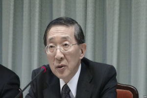 Ein Mann im Anzug und Krawatte sitzt an einem Tisch mit einem Mikrofon, trägt eine Brille und hat eine andere Person Hand auf der linken Seite und einen Vorhang im Hintergrund.