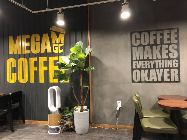 Außenansicht eines Kaffees namens "Mega Coffee" mit Tischen, Stühlen, einem Bodenständer, Deckenleuchten und einem Topfpflanze in Singapur.