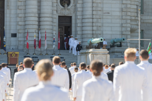 Eine Gruppe von Menschen in weißen Uniformen und Mützen steht vor einem Gebäude mit Säulen und einer Tür, wobei Flaggen, ein Podium mit Mikrofon, Stufen und Kanonen im Hintergrund zu sehen sind, was auf eine Abschlussfeier an einer Marineschule hindeutet.
