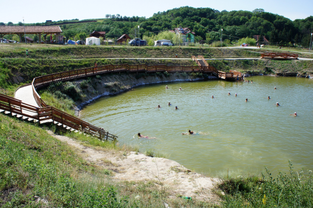 Gruppe von Menschen, die in einem Gewässer schwimmen, umgeben von Grünflächen, einer Brücke mit Geländer und Treppen, Hütten, Fahrzeugen, Pfählen und einem klaren blauen Himmel im Hintergrund.