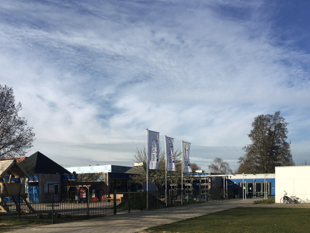 Großes Gebäude mit Metallzaun, Rasenfläche, Weg, Bäumen, Pfosten, Bannern, Fahrrad, nahen Häusern und bewölktem Himmel, als neuer Spielbereich für Kinder an der Schule identifiziert.