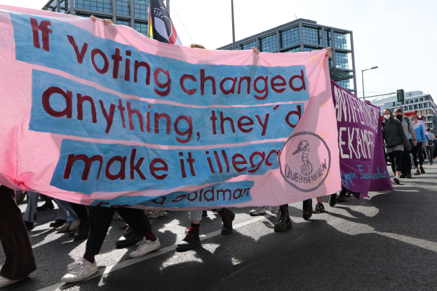 Eine Gruppe von Menschen marschiert mit einem Banner mit der Aufschrift "Wenn Abstimmen etwas ändern würde, hätten sie es verboten" eine Straße entlang, mit Gebäuden, Laternenmasten und einem klaren blauen Himmel im Hintergrund.
