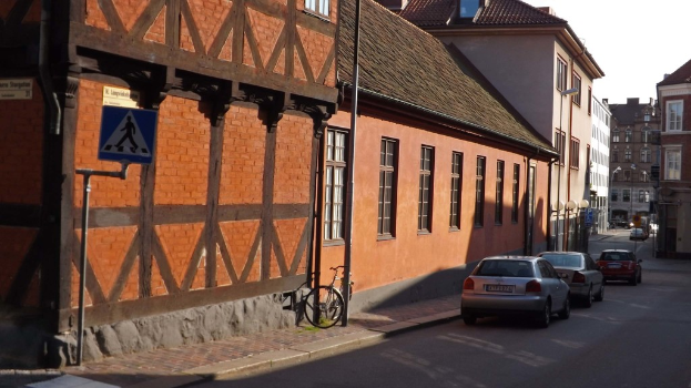 Fahrzeuge auf der Straße mit Gebäuden, Laternen, Verkehrszeichenmasten, Fenstern und Dächern im Hintergrund und einem Fahrrad auf dem Gehweg in der Nähe eines Mastes.