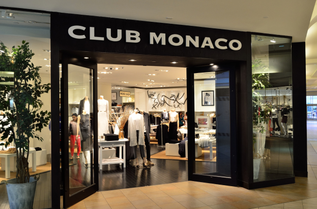 Ein Club Monaco-Laden in einem Einkaufszentrum mit Glaswänden und -türen, der Schaufensterpuppen in Kleidern, Tische mit Gegenständen, einen Topf mit einer Pflanze und Deckenleuchten zeigt.