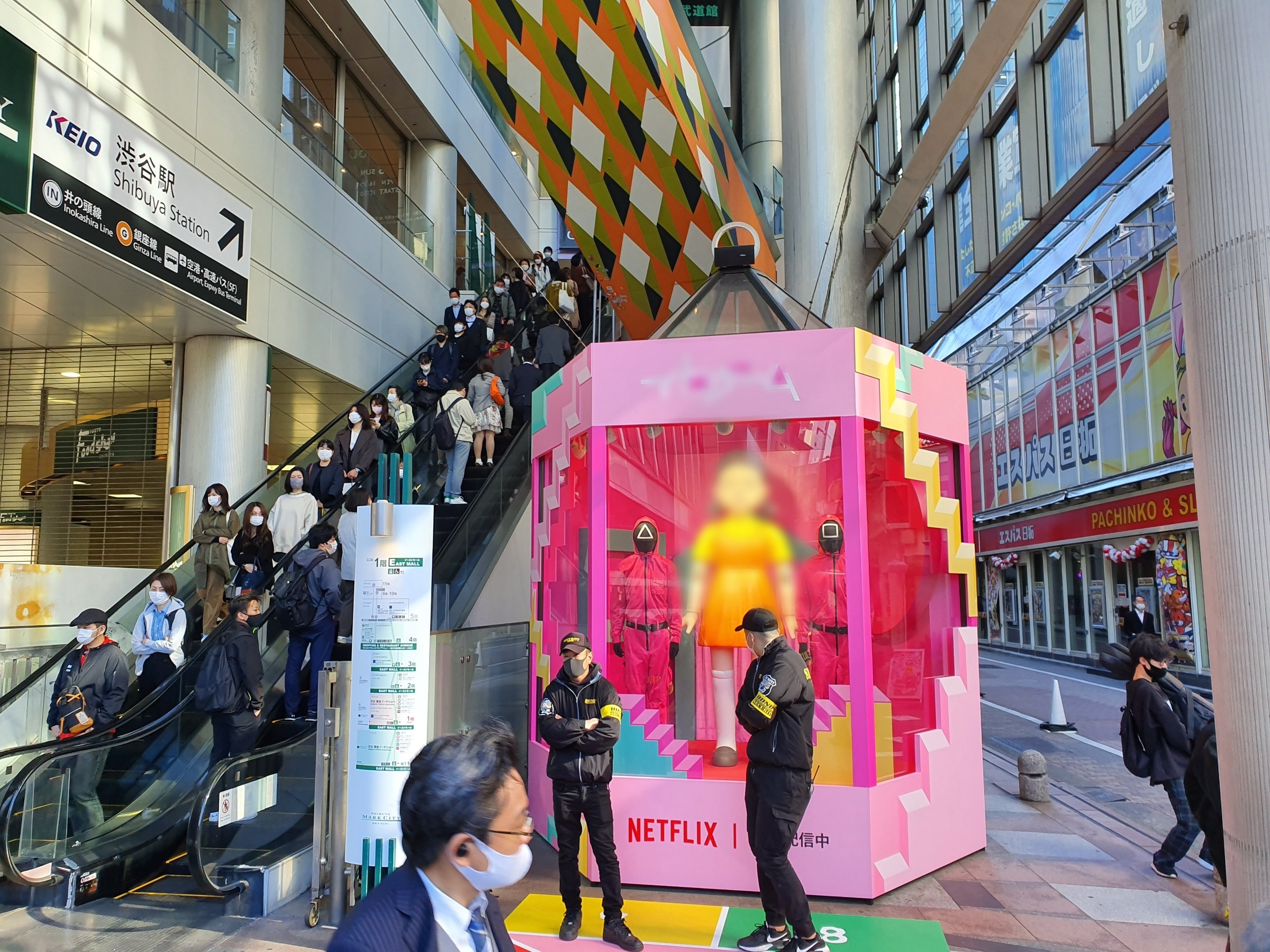 Eine Gruppe von Menschen steht vor einem Gebäude mit Glaswänden, einige tragen Masken, während eine Rolltreppe mit Menschen im Hintergrund zu sehen ist; ein pinkfarbener Kasten in der Mitte zeigt Schaufensterpuppen und trägt die Aufschrift "Netflix's new Netflix booth in Tokyo."