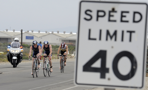 Gruppe von Radfahrern mit Helmen auf einer Straße mit einem Geschwindigkeitsbegrenzungsschild, Gebäude und Bäume im Hintergrund unter einem klaren blauen Himmel.