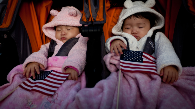 Zwei Babys schlafen in einem orange-schwarzen Babywagen mit weißen, roten und blauen Flaggen im Hintergrund und pinken Decken unten.