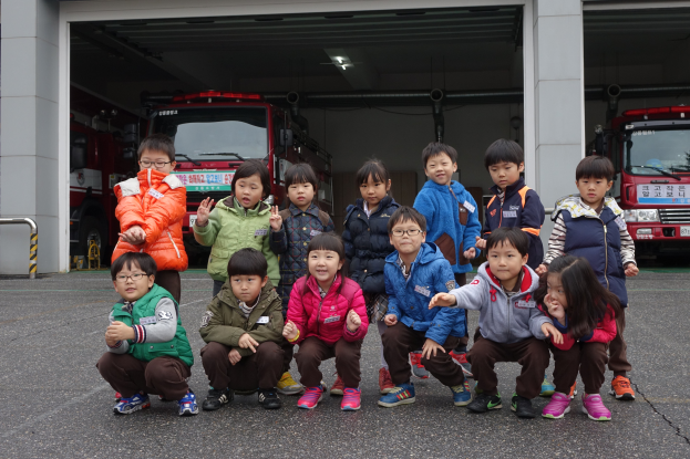 Eine Gruppe von Kindern in Jacken, einige mit Brille, posiert vor einer Feuerwache mit mehreren Feuerwehrautos im Hintergrund und einem Geländer auf der linken Seite.
