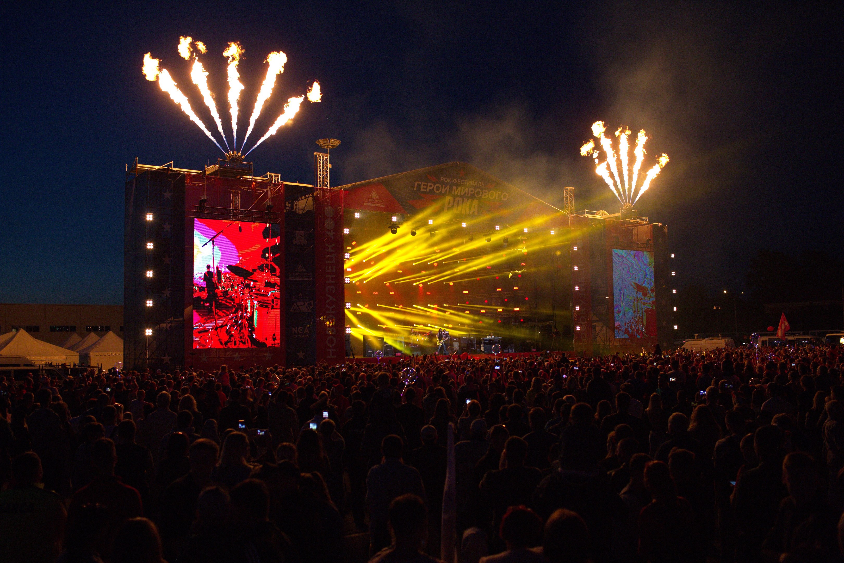 Eine große Menschenmenge auf einem Musikfestival mit beleuchteter Bühne, Performern, Bannern, Zelten links und Feuerwerk am dunklen Himmel.