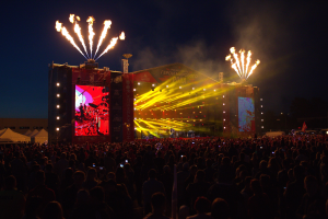 Eine große Menschenmenge auf einem Musikfestival mit beleuchteter Bühne, Performern, Bannern, Zelten links und Feuerwerk am dunklen Himmel.