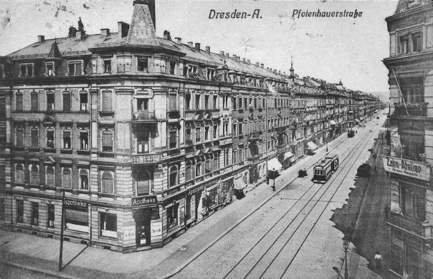 Ein altes Schwarz-Weiß-Foto einer Stadtstraße in Dresden, Deutschland, das Fahrzeuge, Fußgänger, Gebäude mit Fenstern und Schildern, Strommasten mit Drähten und einen Himmel im Hintergrund zeigt, mit Text oben und unten auf dem Bild.