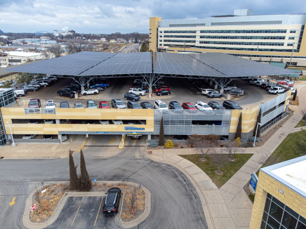 Luftaufnahme eines solarpanelbedeckten Parkplatzes mit parkenden Autos, umgeben von Gebäuden, Grünflächen, Straßen und Hügeln unter einem bewölkten Himmel.