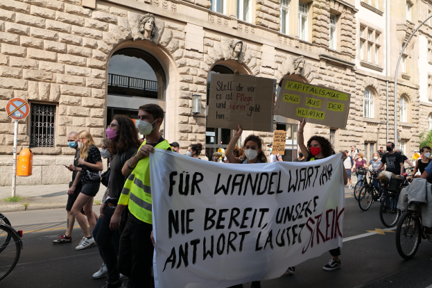 Eine Gruppe von Menschen marschiert auf einer Straße, einige halten Schilder und andere fahren Fahrräder, im Hintergrund ein historisches Gebäude mit Rundbogenfenstern, Säulen und Skulpturen.