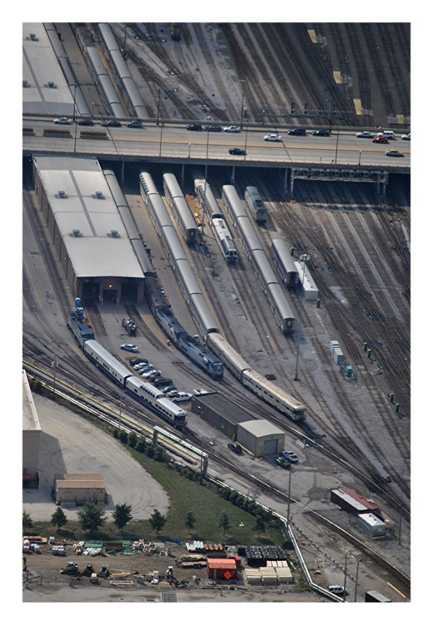 Eine Brücke mit wenigen Fahrzeugen darauf, darunter Eisenbahnschienen mit ein paar Zügen und verschiedenen Gebäuden, Bäumen, Pfählen, Gegenständen und Fahrzeugen auf dem Boden.