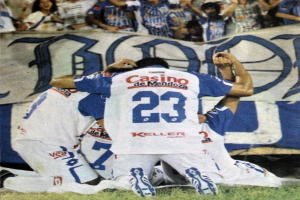 Gruppe von Füßtballspielern, die auf dem Boden vor einer jubelnden Menge mit einem Banner im Hintergrund feiern.