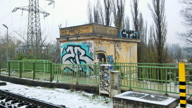 Ein Bahngeleis mit Graffiti an der Seite, umgeben von Gelöndern, Pfosten, Lichtern und Bäumen, mit Törmen und Drähten im Hintergrund und Schnee am Boden.