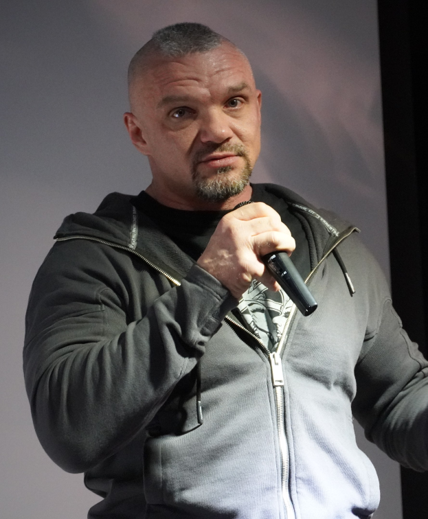 Schwarzkleidiger Komiker in weißem Hemd und zurückgekämmtem Haar hält ein Mikrofon und steht selbstbewusst vor einer Wand.