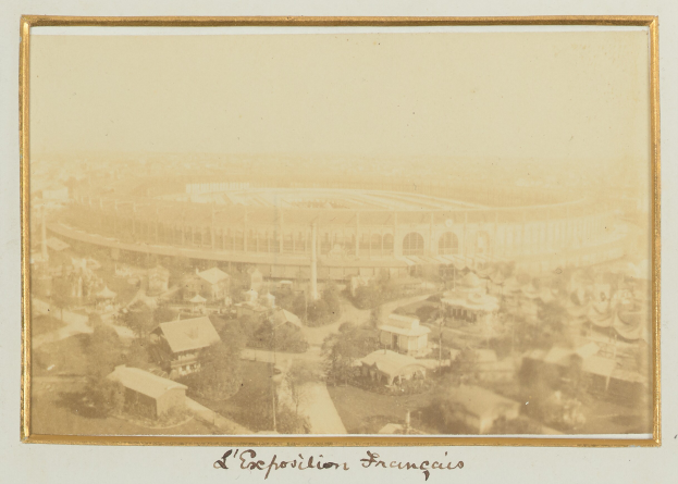 Schwarz-weiß-Foto eines Stadions in städtischer Umgebung, umgeben von Gebäuden, Bäumen und Strommasten, mit Text unten.
