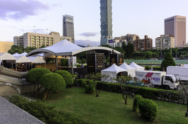 Eine Gruppe von Zelten in einem Park mit Grünfläche, Menschen, Fahrzeugen, Gebäuden und einem klaren Himmel, wahrscheinlich aufgenommen auf dem Hong Kong International Film Festival.